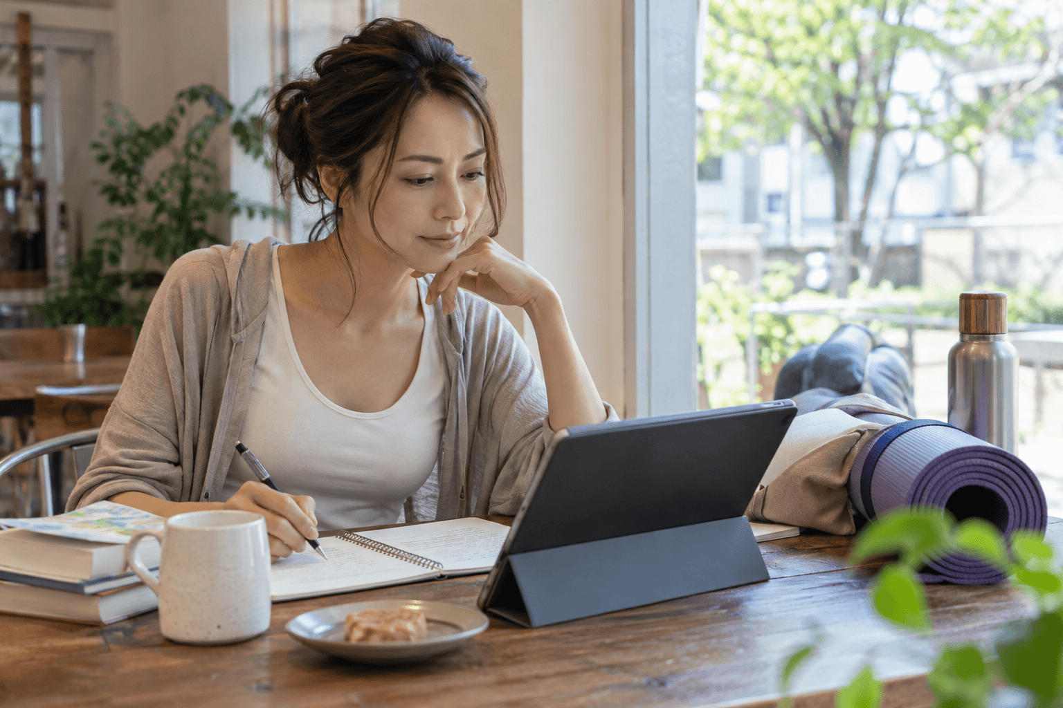 仕事の合間にヨガ資格の学習をする女性。ノートとヨガマットを広げた机でタブレットを見ている